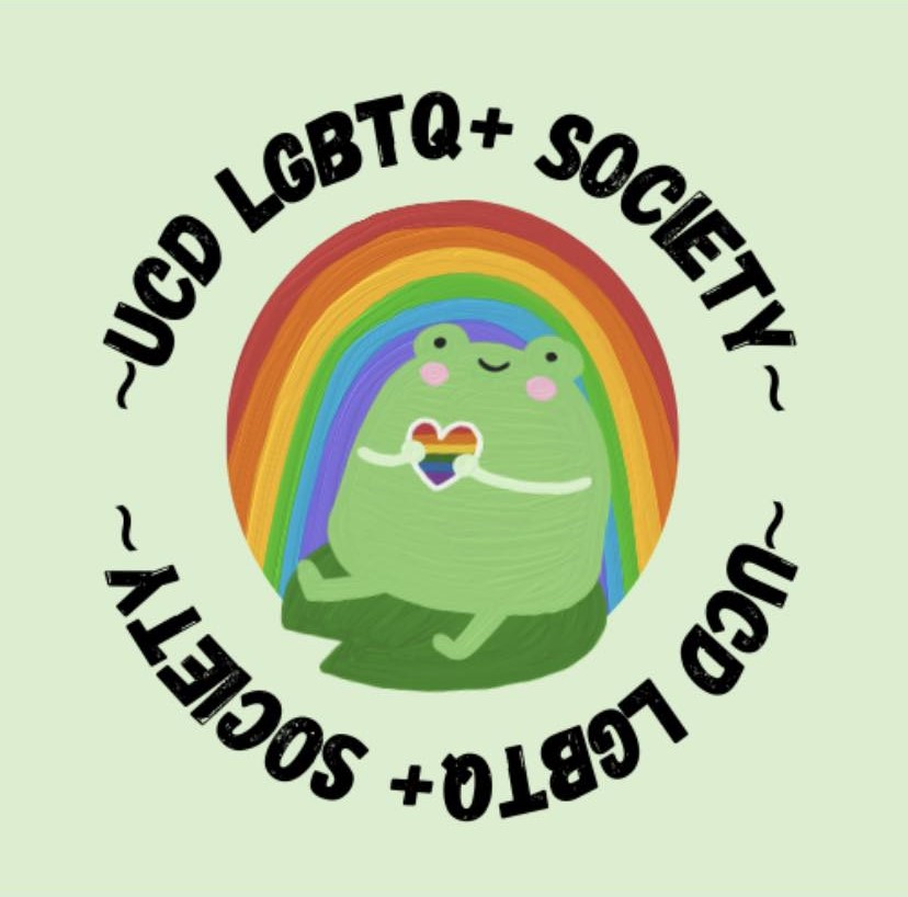 A picture of a green frog holding a rainbow love heart, sitting on a green lily pad, in a leant back position. The frog has two beady eyes either side of its head, a u-shaped smile near the top of its head, and two rosy cheeks underneath the eyes. The frog is sitting in front of a rainbow. The phrase "-UCD LGBTQ+ Society-" is visible above the rainbow, arching around it. The same text is visible underneath the lily pad, this time being upside down.